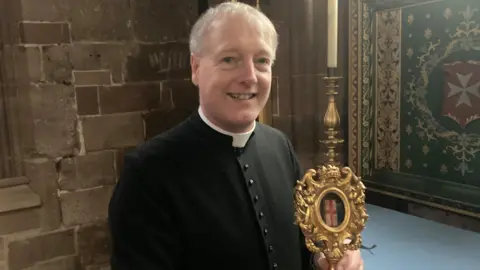 Father Dexter Bracey is wearing a black tunic with a white dog collar and holding a golden reliquary showing a small white oblong box, tied with a red ribbon, that contains a shard of bone purported to be from St Valentine.