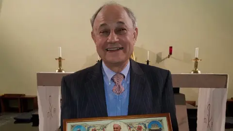 BBC Edgar Riches in a dark jacket with light blue shirt and jumper, standing in front of a church altar.