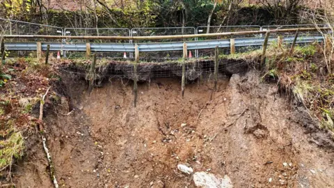 Westmorland and Furness Council The landslip under the road. The road is bordered by a barriers and there are orange cones by the fence too. Underneath the fence is exposed soil.
