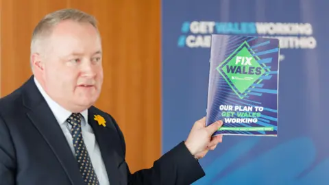 Athena Pictures A man with white hair in a navy suit holds up a copy of pamphlet which reads "Fix Wales".