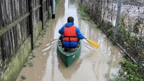 A man with his back to the camera, travelling through floodwater. He is wearing a life jacket, and blue coat. His canoe is green and his oars are yellow. 