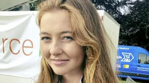 Family handout A close up image of Katie Simpson smiling at the camera. She has long brown hair with blonde highlights and is standing in front of a banner, with unreadable text. 