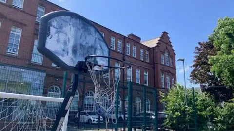 A sad looking basketball hoop with the corner of a white football goal behind it. Westgate Hill Primary Academy is behind it which is a large red brick Victorian building.