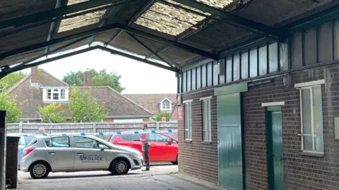 A silver police vehicle is parked at the scene where the worker fell, with the area cordoned off. The image shows a roof over an empty area, which has an open end. The right side has doors and windows of a building.