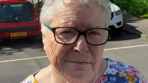 Carol has short grey hair and black rimmed glasses and is wearing a floral top.