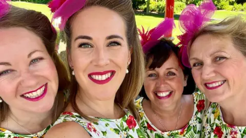 Selfie of four smiling women in matching clothes and make-up. They are wearing sleeveless floral dresses, dark pink lipstick and cerise fascinators.