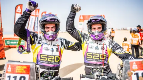 Craig and Carl Searles Two men in motorcyle gear and purple helmets are cheering, while sitting on their machines. Behind them is the desert, with series of banners for the rally, and photo-accredited people to the right of the shot. 