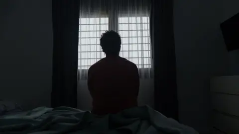 A silhouette of the back of a man sitting on the edge of a bed looking out of a window.