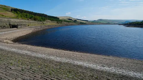 Exposed stone and bricks are revealed at Woodhead Reservoir, where the water level has lowered significantly. The water is surrounded by green hills, covered in trees.