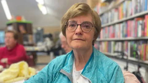 An older woman with short, light brown hair. She is wearing glasses, a white t-shirt and a blue zip-up top. She is sitting in a library, with a row of books visible behind her.
