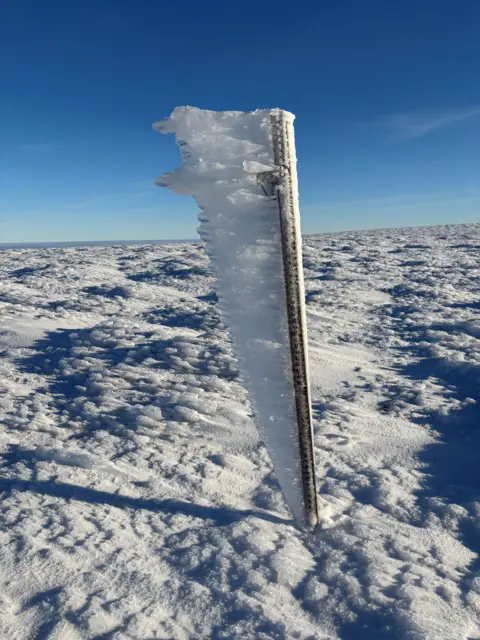 Peter Braidwood Metal pole sticking out of snow covered ground with ice coating one side of it as the wind has blown it in one direction