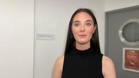 Kate has long dark hair is standing indoors against a white wall. She is wearing a sleeveless black top. In the background, there is a grey door with a circular window on the right, and a sign on the left that reads 'PLEASE DO NOT TOUCH'.