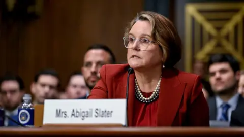A woman wearing a red suit sits in front of a microphone.