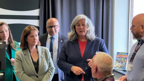 A woman in a navy blazer and long grey hair speaking to a man sat down in front of a computer 