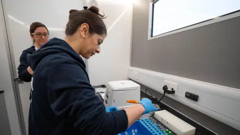 NIHR A research nurse handling some test tubes in the back of a van with a colleague beside her.