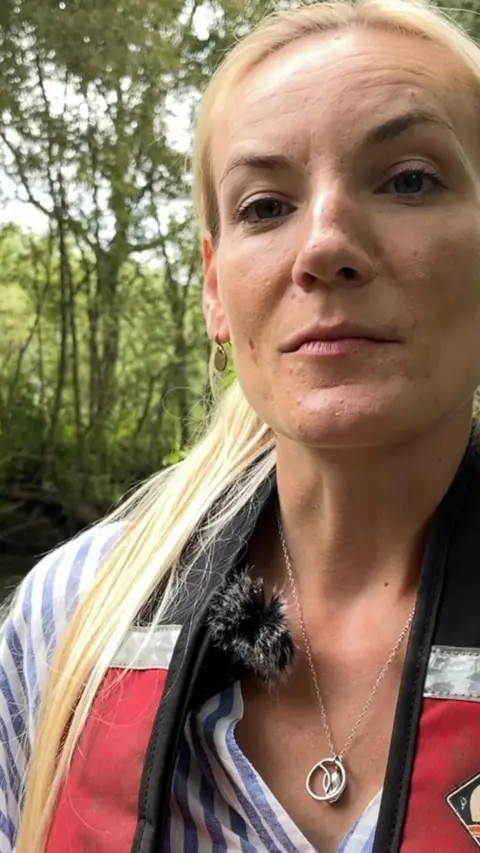 A woman wearing a life jacket is standing in a stream, she has blonde hair.