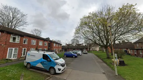 A cul-de-sac with two-storey red brick buildings and cars parked on the left side and trees on the right.