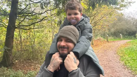 Family A man with a short beard wearing a grey beanie smiles towards the camera. His young son is sitting on his shoulders, he is wearing a grey coat and has a purple dummy in his mouth. They appear to be in a park or on a woodland trail, there are trees and a dirt path behind them