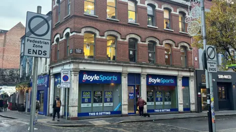 Two grey and white signs mark the end of a pedestrian zone in Belfast. There is a Boyle sports shop in the background. Above the shop a red brick building continues. There is a cobbled street and footpaths between the signs and the building.