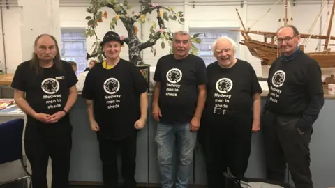 Medway Men In Sheds Five men pose for a photograph wearing black Medway Men In Sheds t-shirts in their workshop. A wooden model of a sailing ship is behind them.
