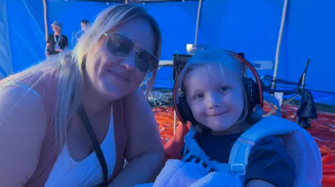 BBC/Jim Scott Lauren Empson-Devonport, wearing a white shirt, is next to her other child, Nova-Leigh, who is wearing headphones. They are both inside the blue sensory zone.