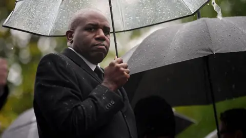 Getty Images David Lammy stands under a clear plastic umbrella and is looking to his right into camera. He is wearing a black raincoat, white shirt and black tie and looks very serious. There are many raindrops on the umbrella. 