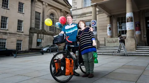 Jonas Pollard Pollard stood next to a woman holding a charity money collector. He is stood next to a bike with three balloons next to him. 