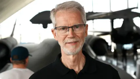 John Fairhall/BBC Steve Justice in a hangar at IWM Duxford. Behind him are two aeroplanes and a vast window. He has white hair and a white beard and is wearing glasses and a black polo shirt. He is just beginning to smile.
