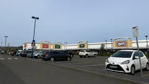 The image shows a retail and leisure park car park on a bright, clear day. The sky is cloudless and light blue, suggesting good weather and daytime conditions. The scene appears calm and orderly, with no visible crowds.