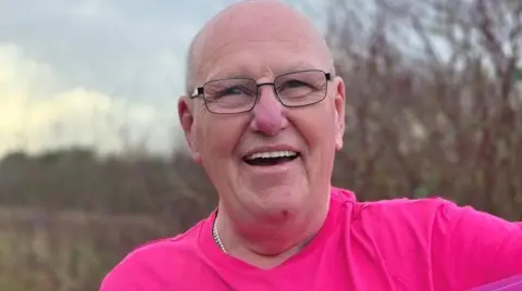 A man wearing glasses and a pink top is looking at the camera. He is outside and bushes and trees are blurred behind him in the photo.