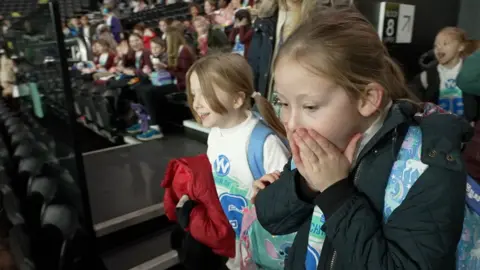 Two school girls in an arena. One has her hands over her mouth in shock