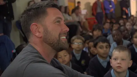 Brown haired man sat down, with kids sat down out of focus in the background