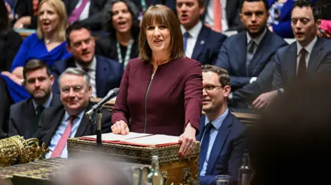 Reuters Rachel Reeves speaking in the House of Commons, with MPs on benches behind her. She has shoulder-length straight dark hair and wears a burgundy dress