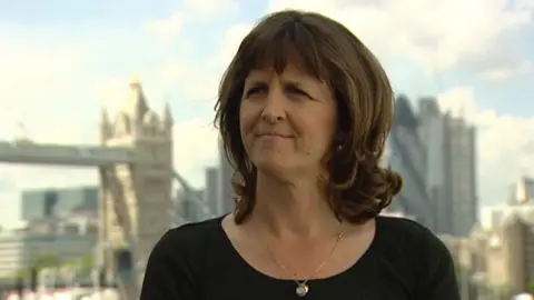 A woman with shoulder-length brown hair is wearing a black top and gold necklace and staring to the left of the camera