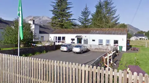 Google A small primary school in the Highlands, a low white building with a couple of cars parked in front