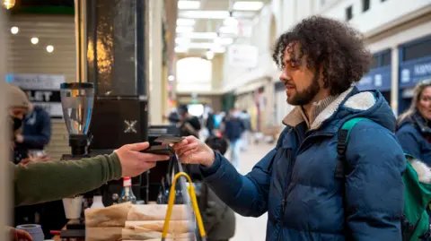 Getty Images Un joven de pelo rizado y chaqueta acolchada azul paga algo en el mostrador de una cafetería con su tarjeta de débito. 