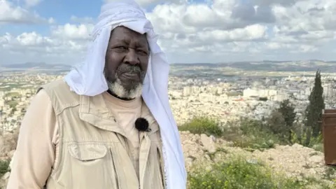 BBC A man with a headdress looks at the camera with a town or city in the background