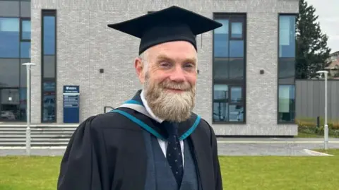 A man wearing a graduation outfit, including the hat and gown, over a suit with a tie. He is standing and smiling outside a university building.