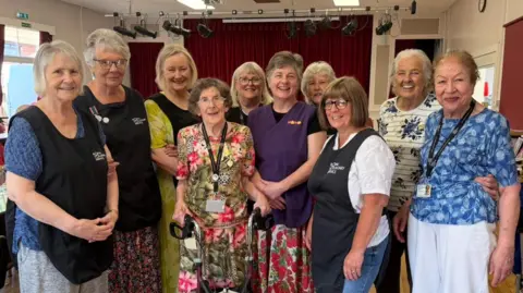 The photo shows a group of women smiling at the camera. Most are wearing black aprons and lanyards. 