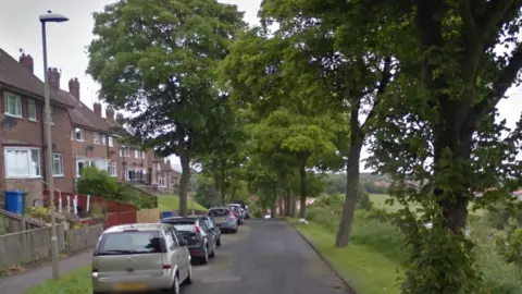 Google A view straight along Barrowcliff Road in Scarborough, with red-brick houses and cars on the left of the road, and trees and fields to the right.