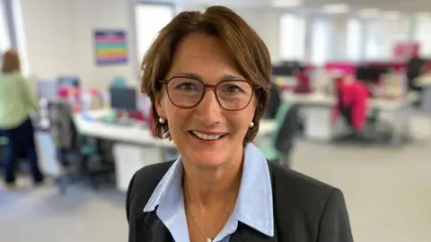 DJ McLaren/BBC Claire McIntosh standing inside an office. She is smiling and looking directly at the camera. She is wearing a blue shirt and a grey blazer.