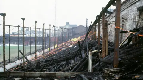 The remains of a football stand following a fire. Charred wood is strewn across the floor while bare, metal stanchions protrude upwards. Workers in yellow protective clothing can be seen at the far end.