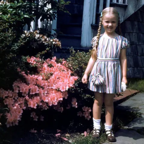 Tom Hodge A child with her blonde hair in plaits is standing in a garden next to a bush covered in pink flowers. She is wearing a summer dress and ankle socks and smiling at the camera. There is a small flight of steps behind her. 