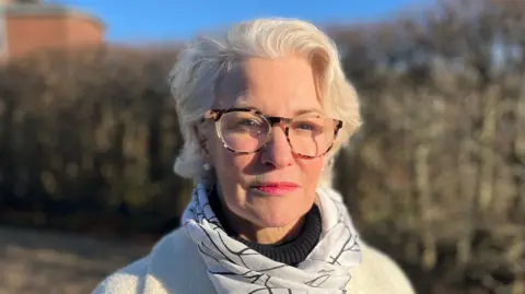 NHS Devon's chief nursing officer Penny Smith stands in front of a large bush on a sunny day. She has white hair and is wearing yellow and brown-rimmed glasses. She is wearing a black and white scarf, a white jumper and black roll neck.