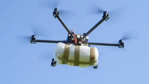 A multi-rotor drone flying in the sky while carrying a package suspended beneath its frame. The photograph is taken from below the drone, looking upward toward a clear blue sky.