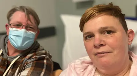 Natalie and her partner Karen are sitting in the emergency department. Karen Davis (left) has short blonde hair and is wearing a checkered brown and cream jacket and a blue face mask. Karen has short brown hair and is wearing a pink panama top.