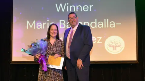 Maria Barandalla-Fernandez wearing a dress and holding a certificate and a bunch of flowers. She is standing in front of a projector with her name on it and the word 'winner'. There is a man in a suit next to her.