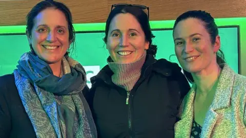 Three women, all with dark hair, stand side by side and are smiling.