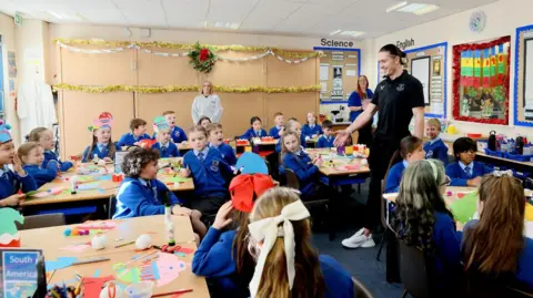 EFC Jack Grealish stand and smailes at the front of a classroom with about 20 children. The children are spread around five big tables with various Christmas cutouts in front of them. The children looked surpised to see him as he moves to shake hands with Georgie at his table. Tinsel and school artwork decorate the walls around the room.