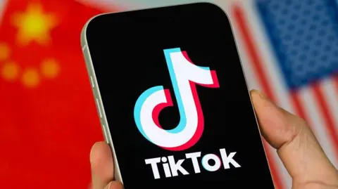 Homescreen of smartphone with TikTok logo, held by a hand, with the flags of China and the US in the background